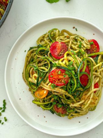 White plate with zucchini pasta topped with halved cherry tomatoes.