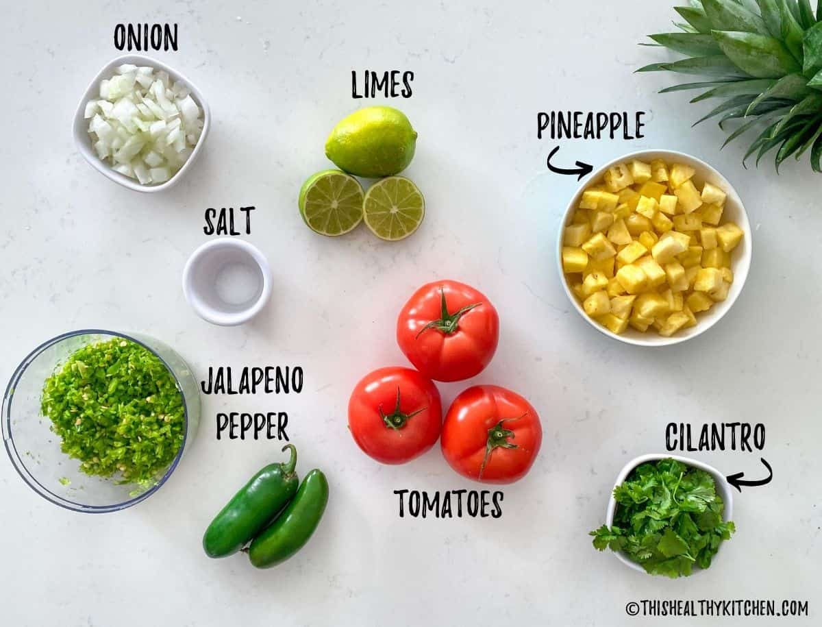 Bowl of pineapple, onion, limes, salt, tomatoes, cilantro and hot peppers on kitchen counter top.