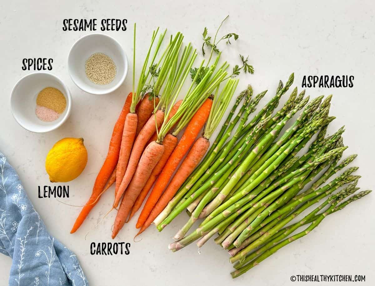 Carrots, asparagus, spices, lemon, and sesame seeds on countertop.