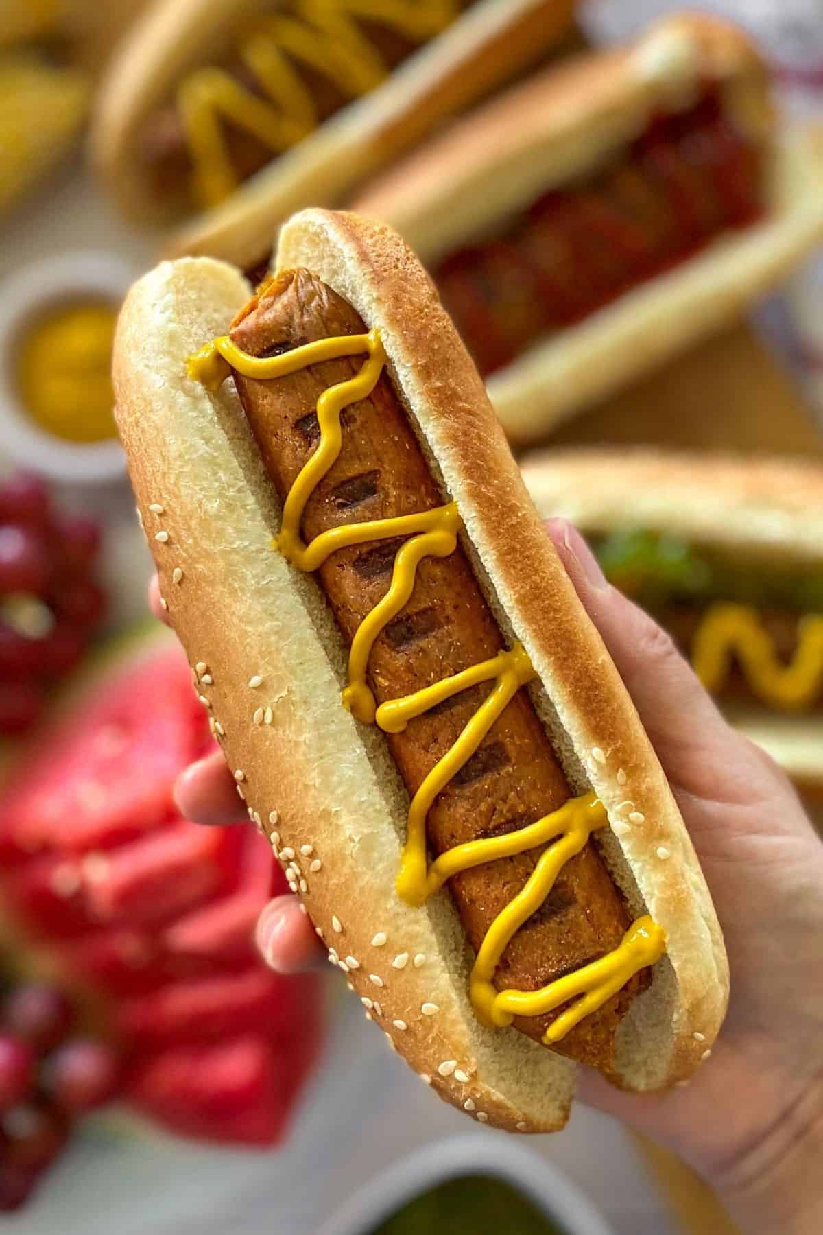 Hand holding up hot dog in bun with mustard on top in zig zag pattern.