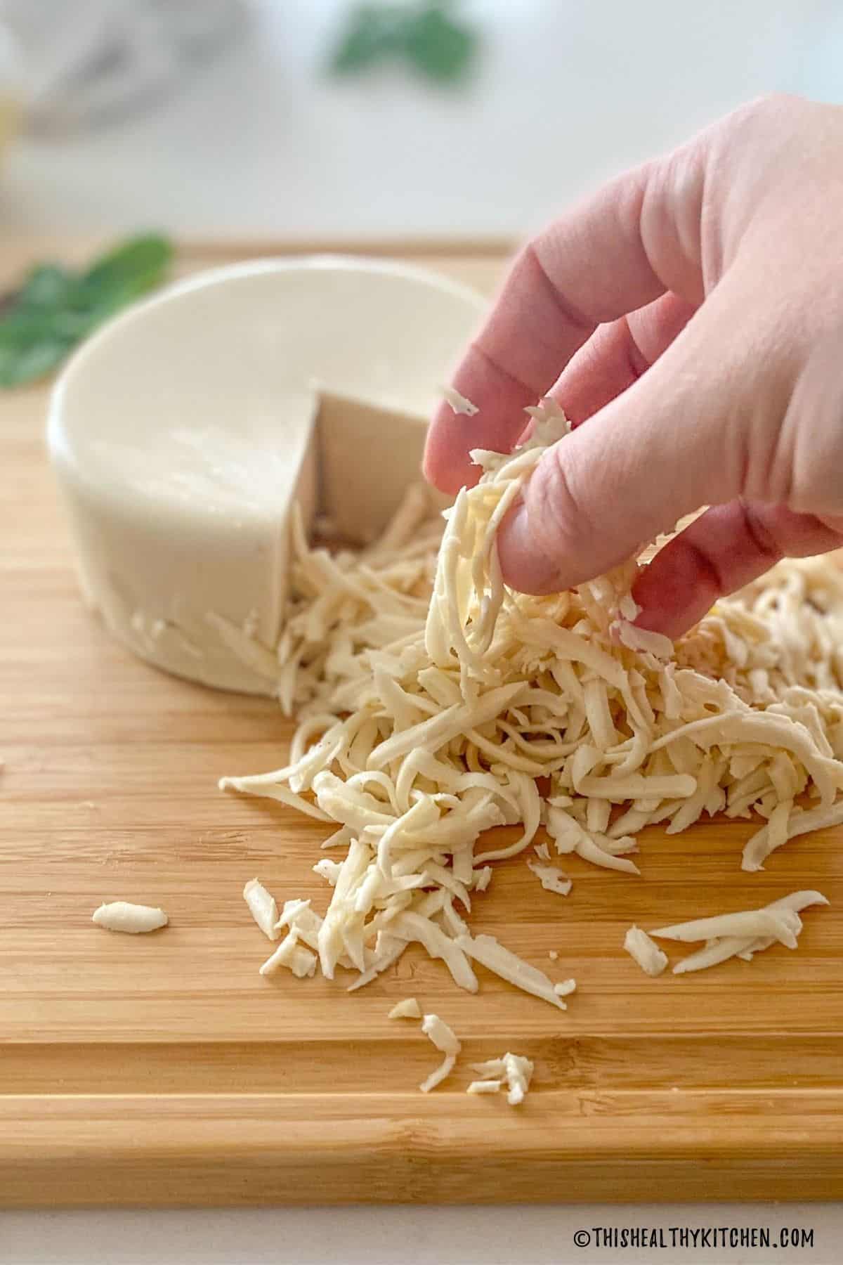 Hand lifting cheese shreds sitting in front of round cheese block.