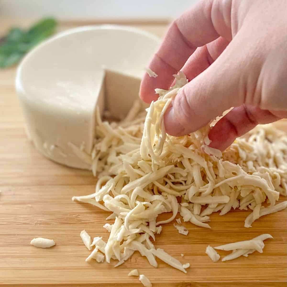 Hand lifting cheese shreds sitting in front of round cheese block.