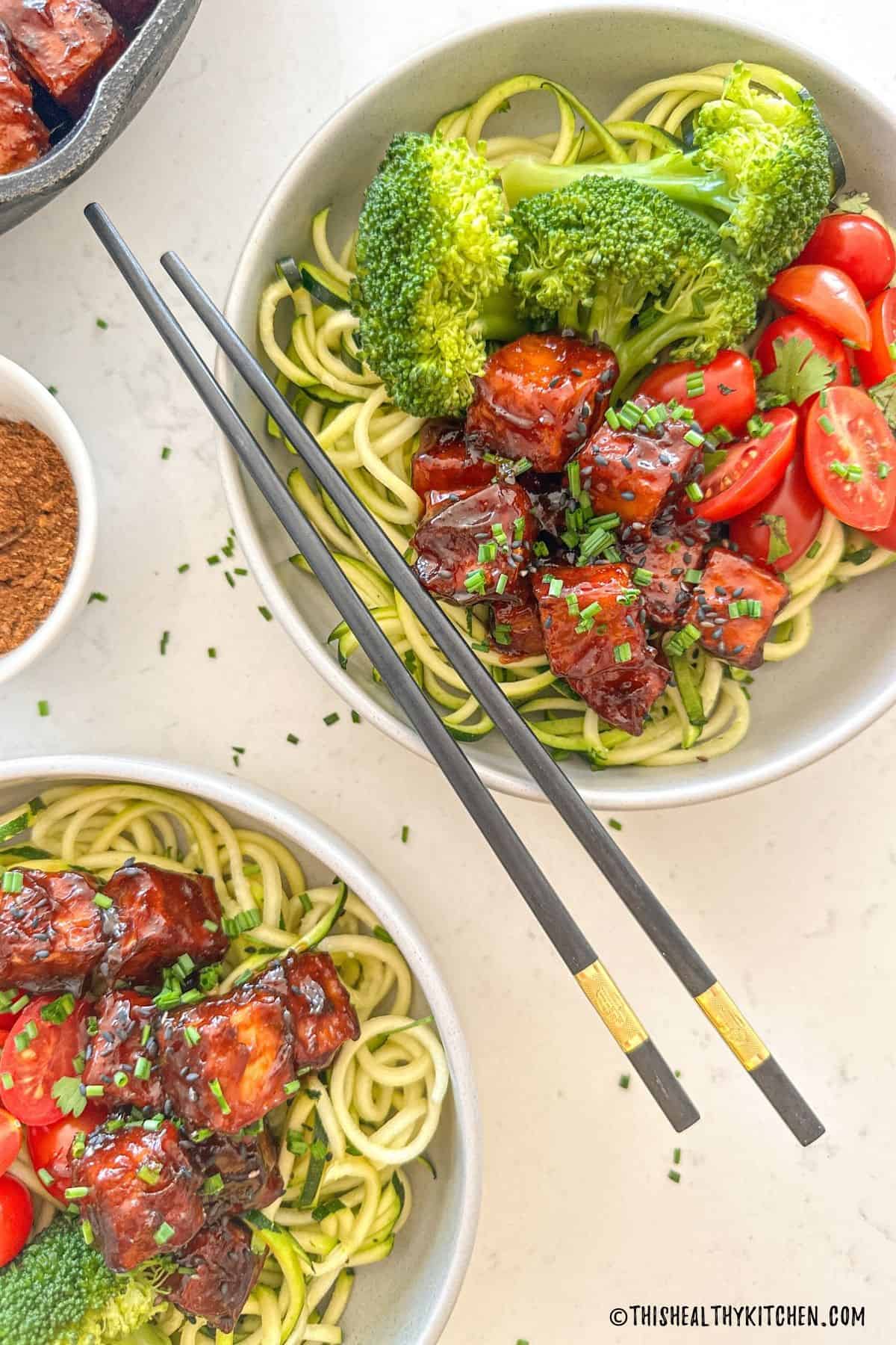 Two bowls of zucchini noodles with broccoli, grape tomatoes and spicy tofu on top.
