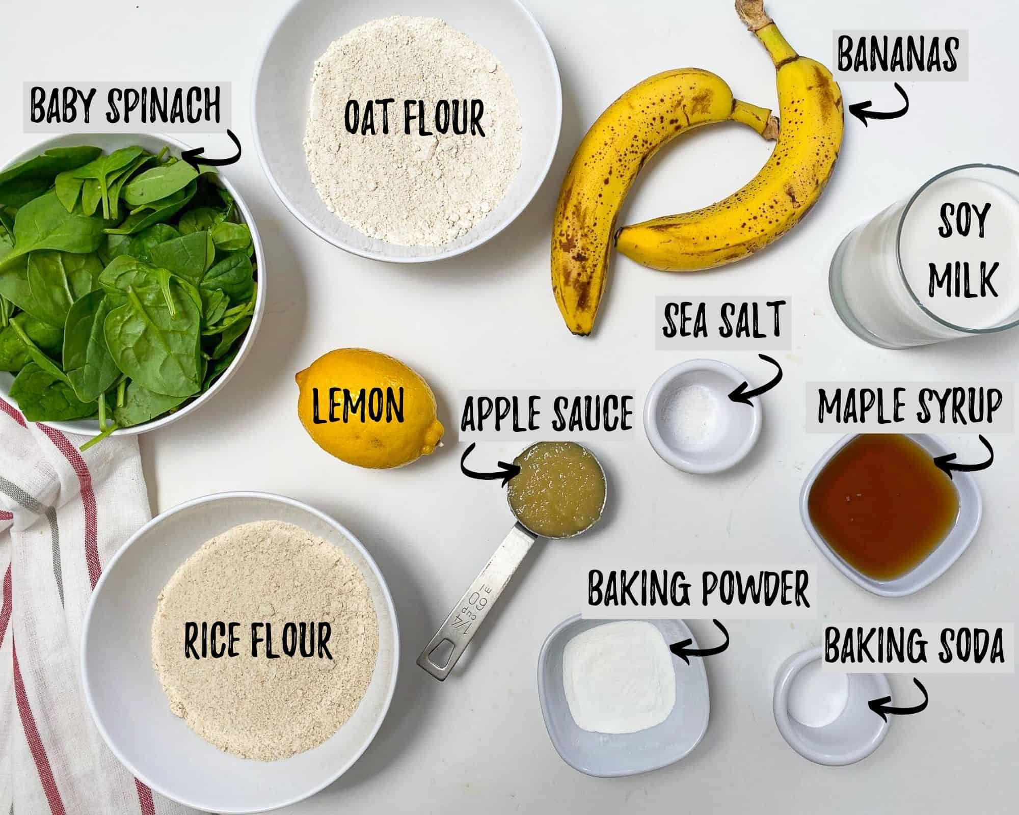 bowl of flour, bowl of spinach, bananas, maple syrup, a lemon, baking powder and soda and applesauce on white counter top
