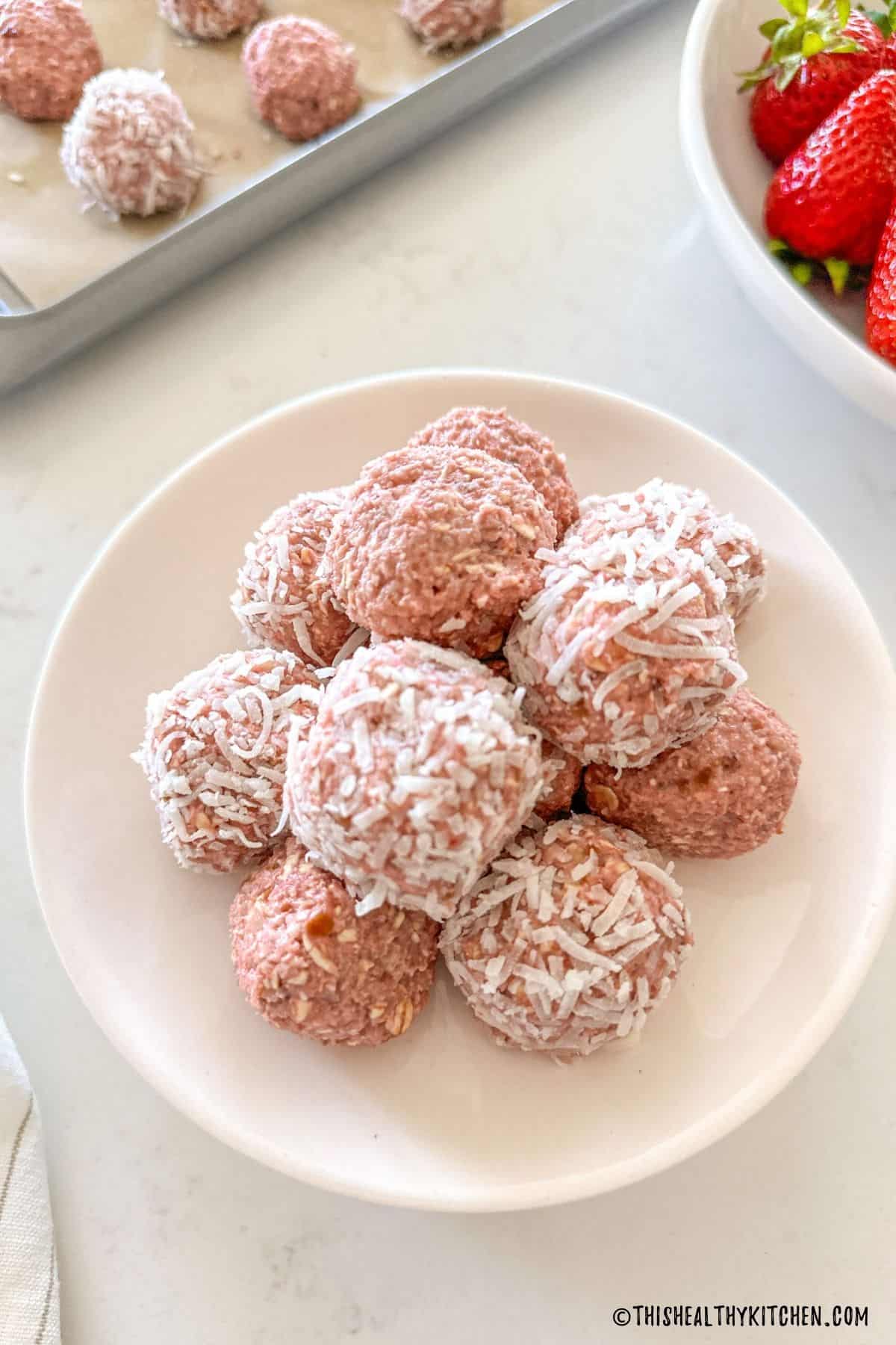 Strawberry bliss balls on white plate with bowl of strawberries in the background.