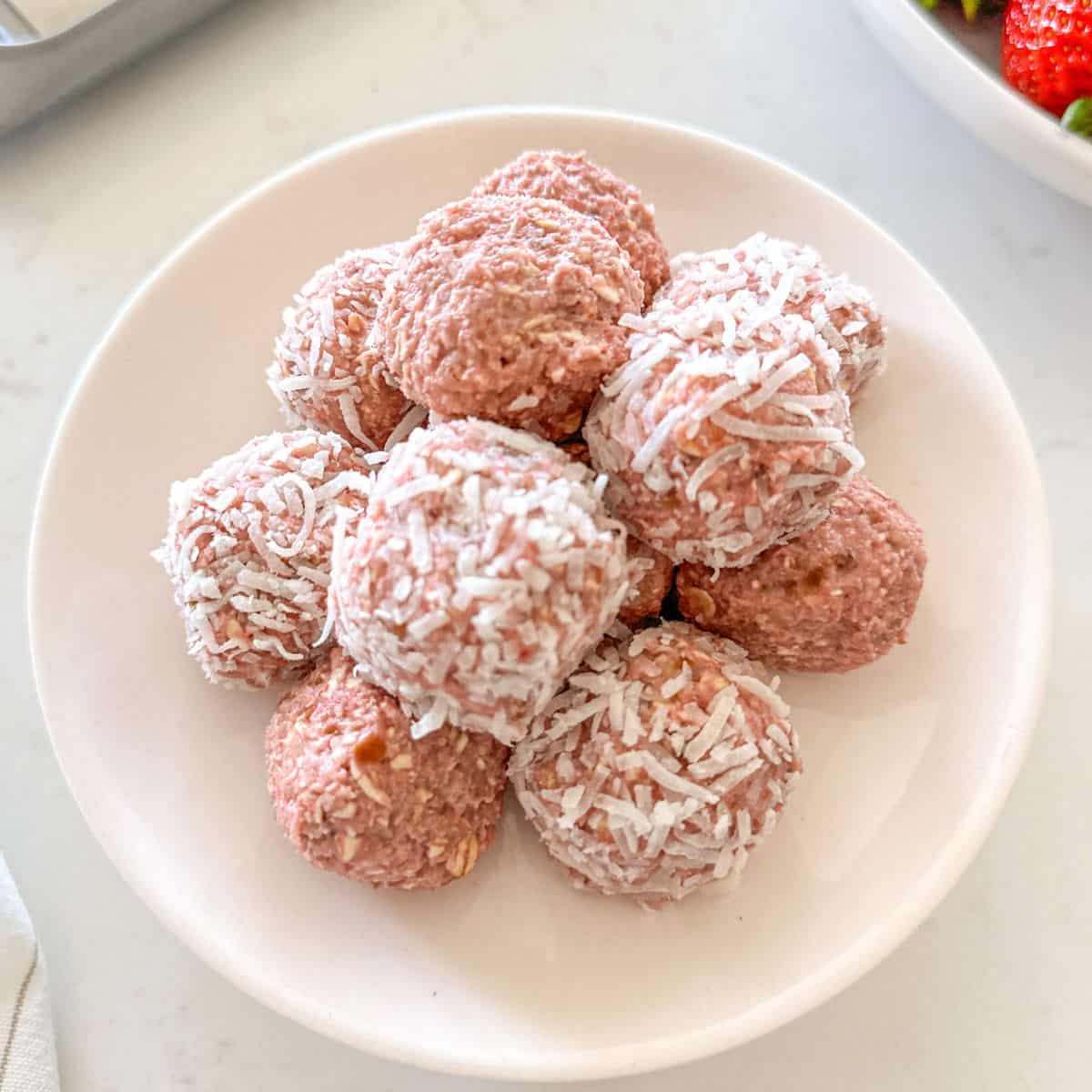 Strawberry bliss balls on white plate with bowl of strawberries in the background.