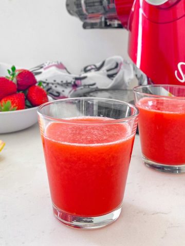 Two glasses of strawberry juice with juicer behind them.