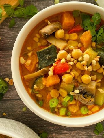 Bowl of soup with sweet potato, mushrooms, zucchini, chickpeas and crushed peanuts and cilantro on top.