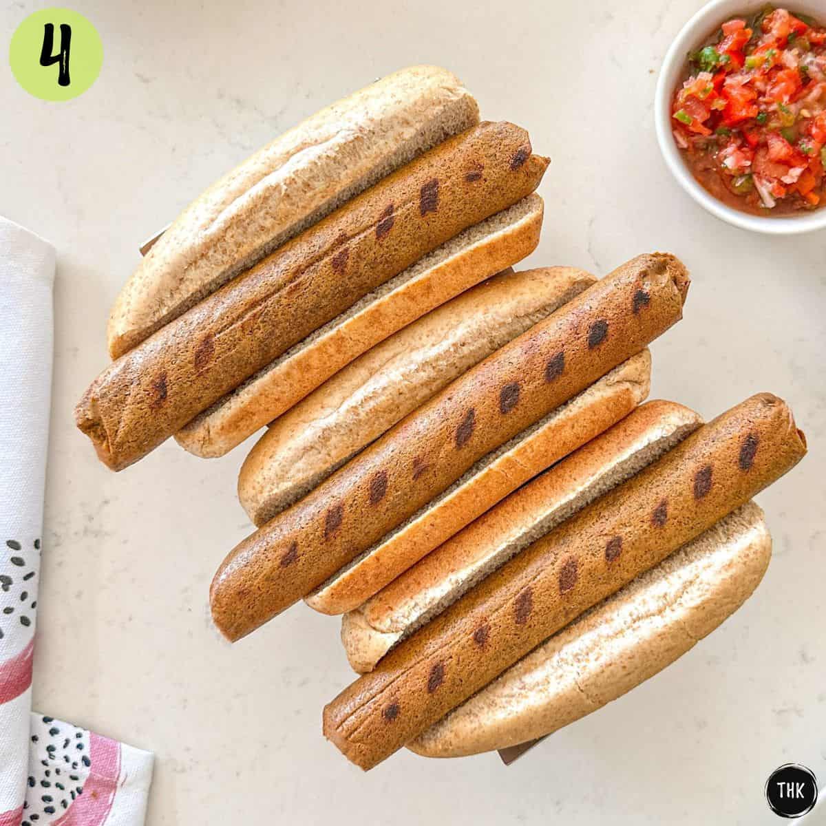 Grilled tofu dogs sitting inside hot dog buns with bowl of tomato salsa on the side.
