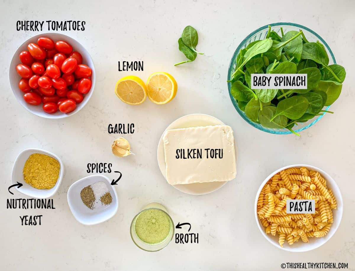 Tofu, pasta, spinach, tomatoes, spices and lemon on kitchen counter top.