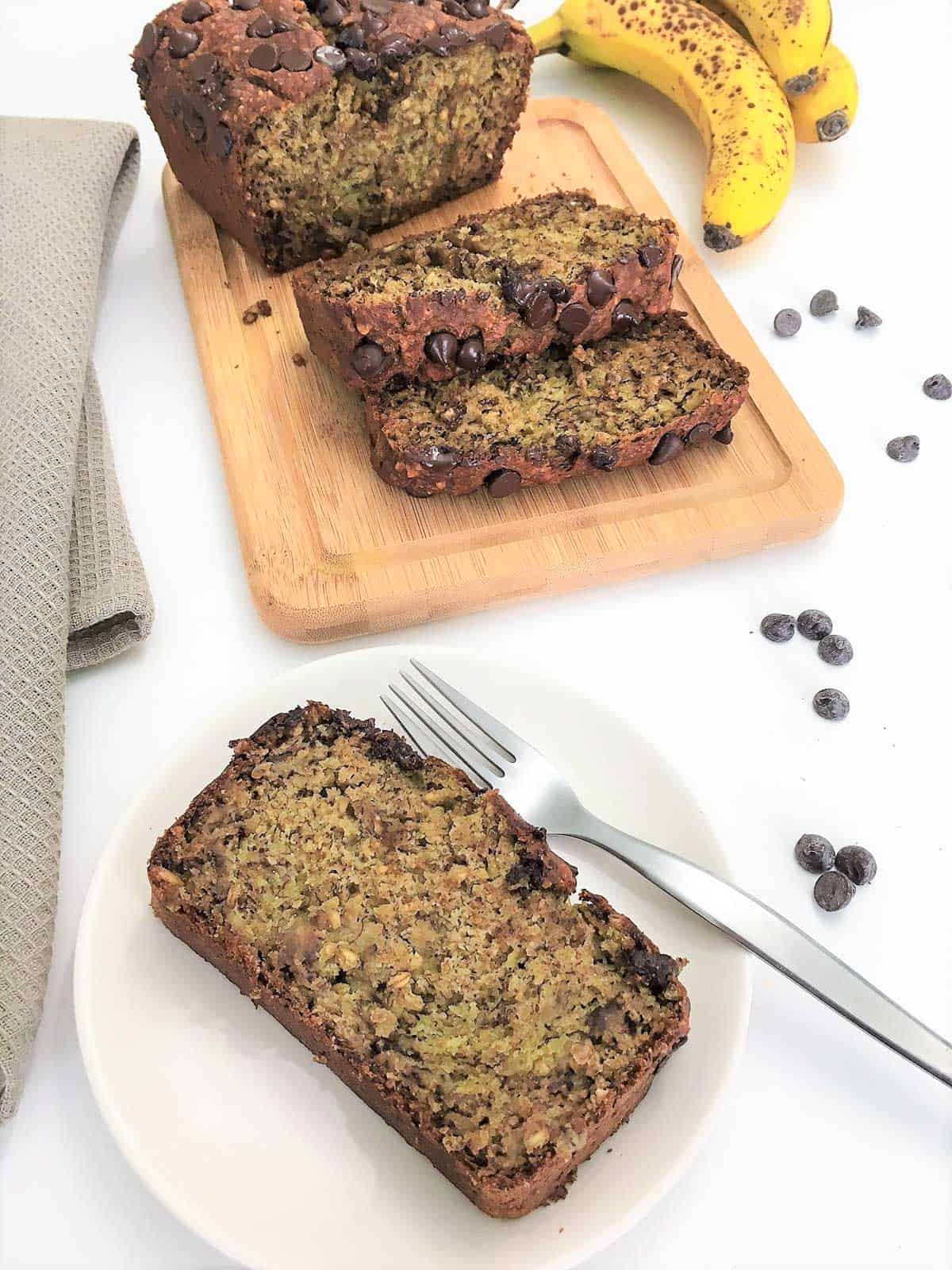 Slice of banana bread on white plate with fork and sliced loaf in background.