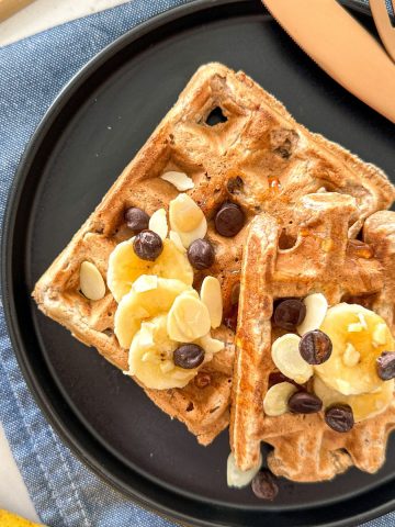 Two banana waffles in black plate with chocolate chips and fork and knife beside it.