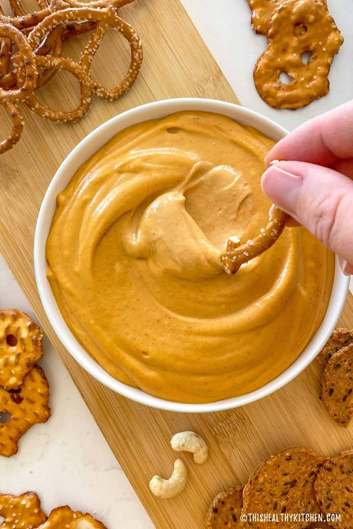 Hand dunking pretzel into bowl of vegan cheese dip.