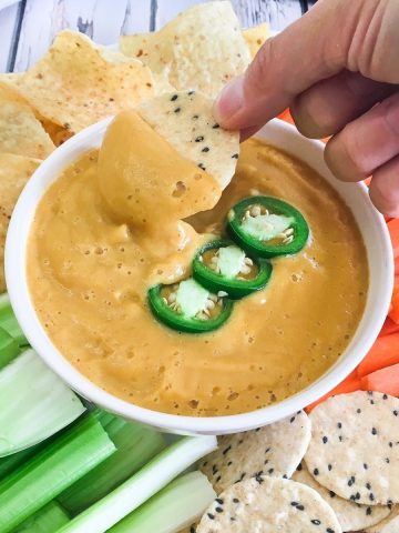 Hand dipping cracker into bowl of vegan cauliflower cheese sauce.