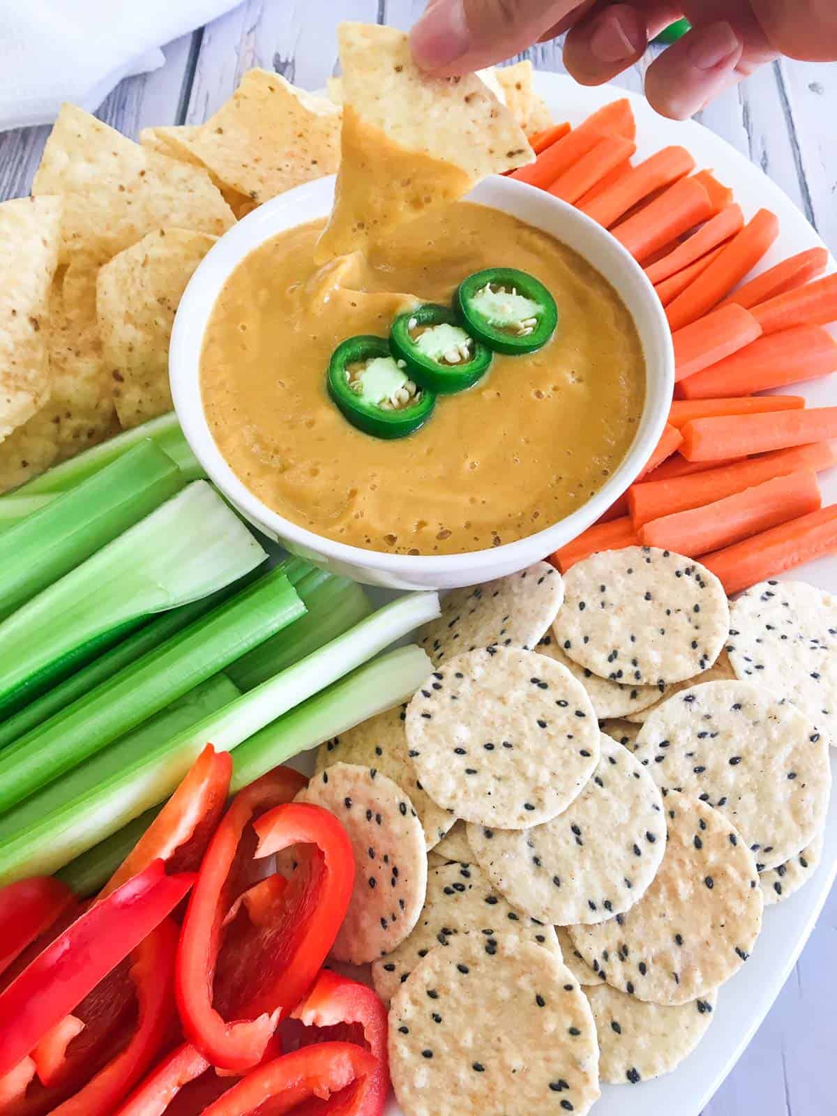 Tortilla chip dunked into cheese dip resting in white bowl with veggies and chips around the bowl.