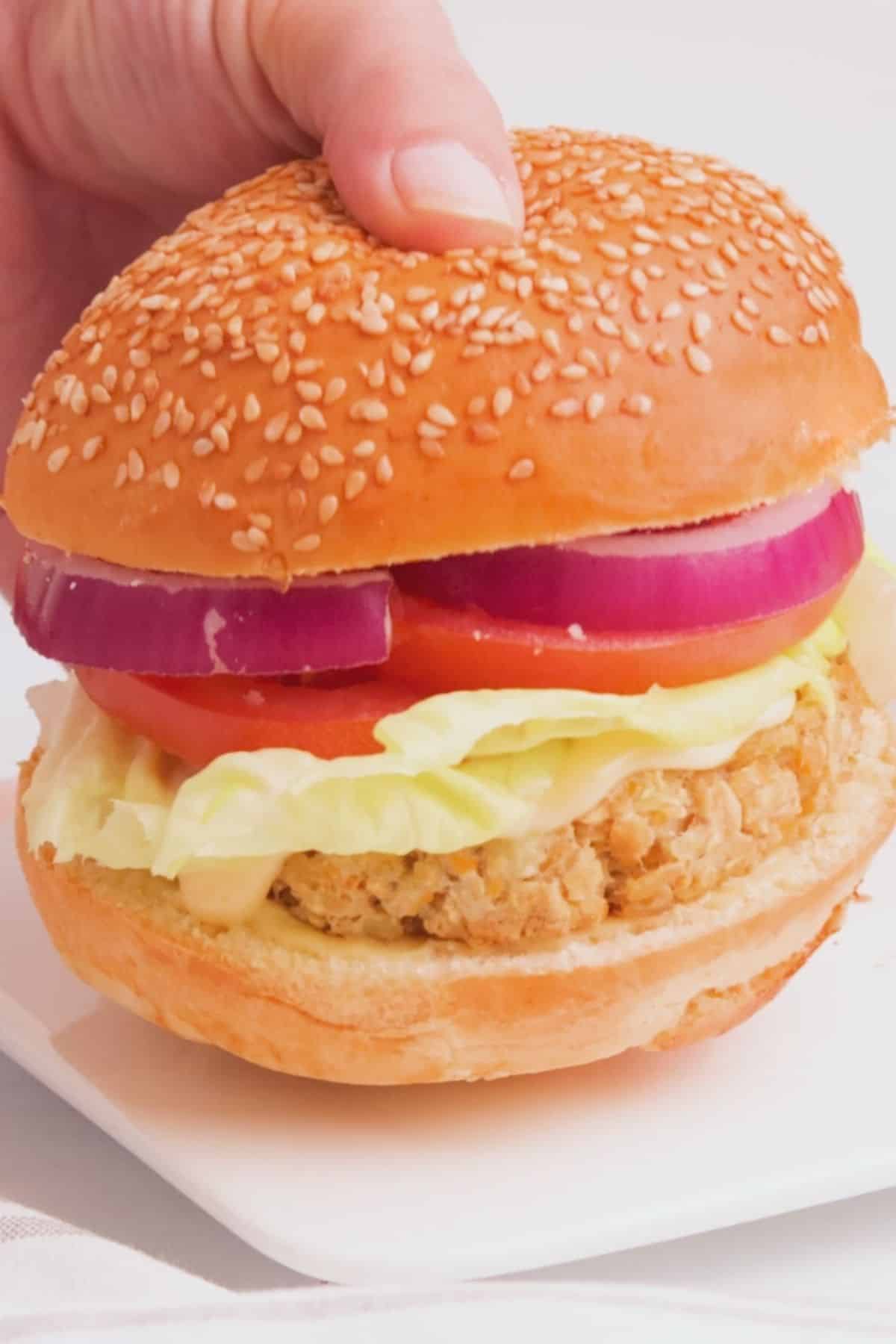 Hand holding up vegan chicken burger with lettuce, tomato and red onion inside.