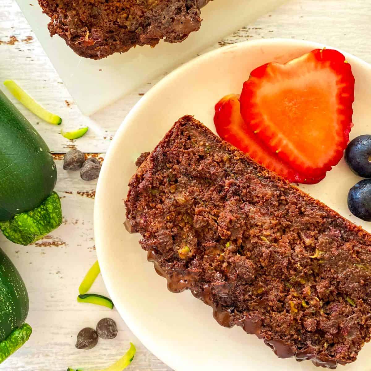 Slice of chocolate zucchini bread in plate with strawberries and blueberries beside it.