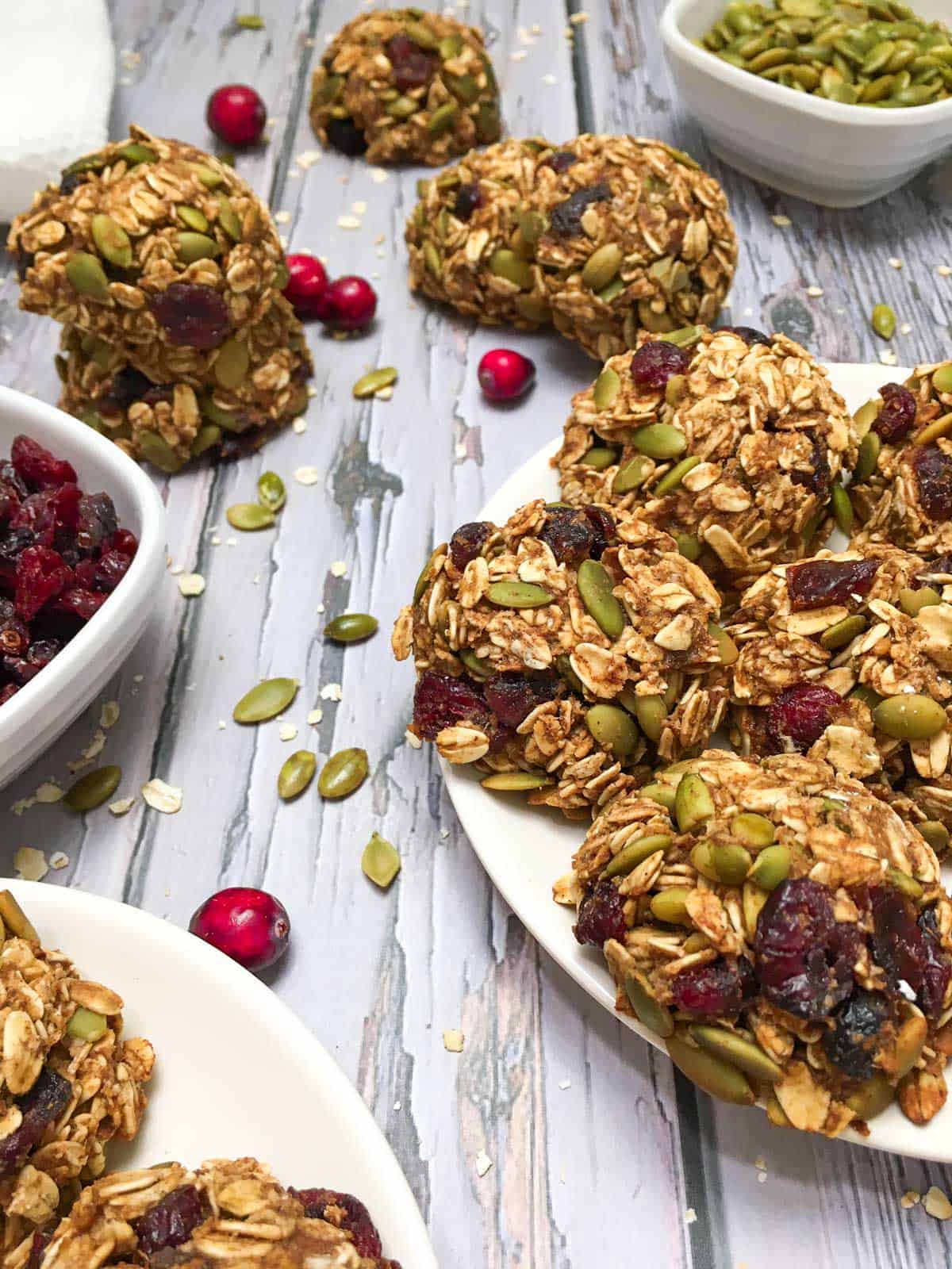 Cranberry oatmeal cookies on white plate with bowls of cranberries and pumpkin seeds around it.