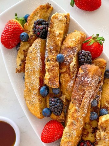 French toast sticks in white platter with strawberries, blueberries and black berries on top.