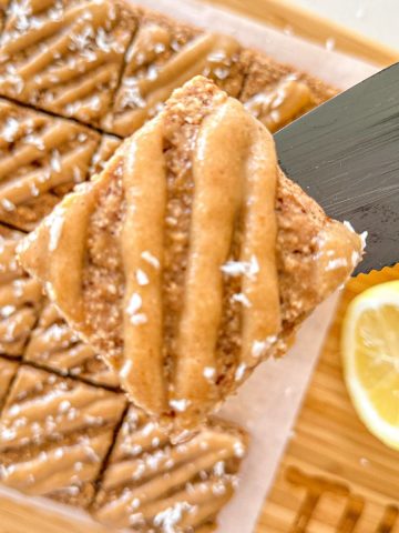 Slice of vegan lemon loaf being held above cutting board with remaining loaf.