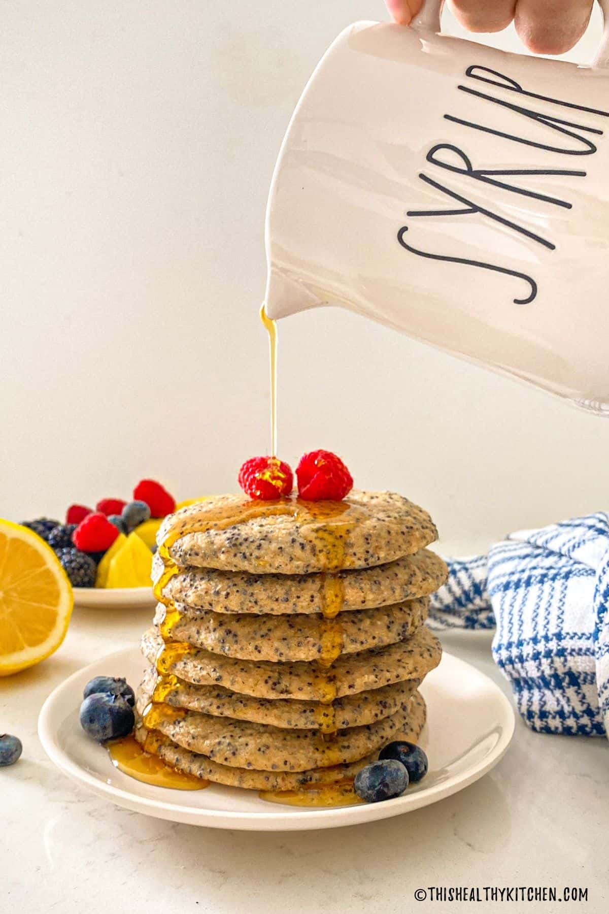 Stack of pancakes on white plate with syrup being poured on top.