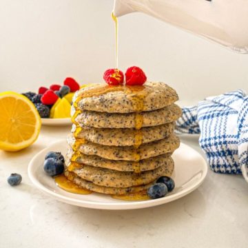 Stack of pancakes on white plate with syrup being poured on top.