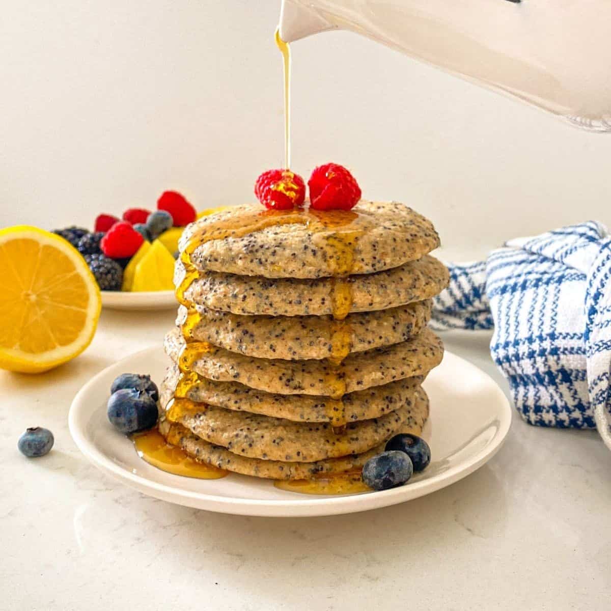 Stack of pancakes on white plate with syrup being poured on top.