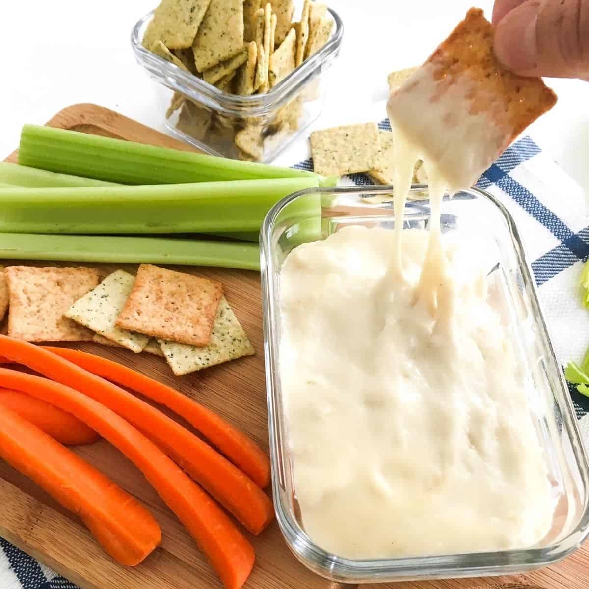 Cracker being dipped into bowl of cheese dip with veggies scattered around it.