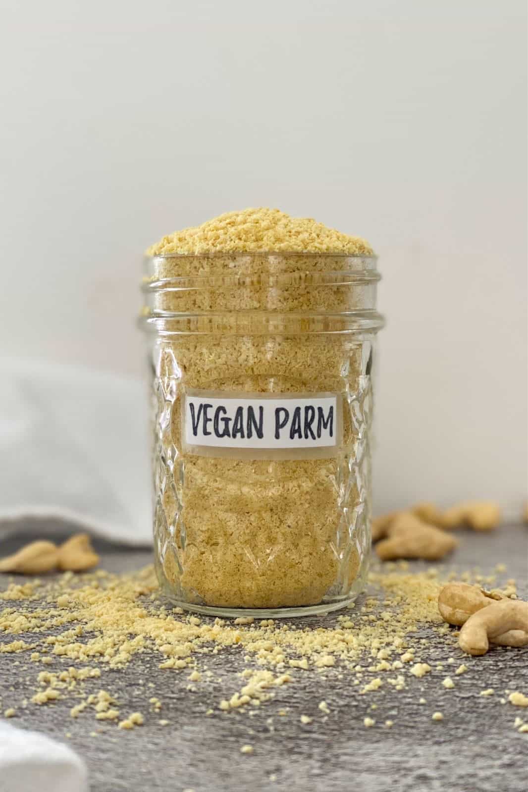 Vegan parmesan cheese overflowing in glass jar with cashews scattered around it.