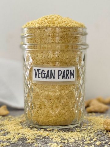 Vegan parmesan cheese overflowing in glass jar with cashews scattered around it.