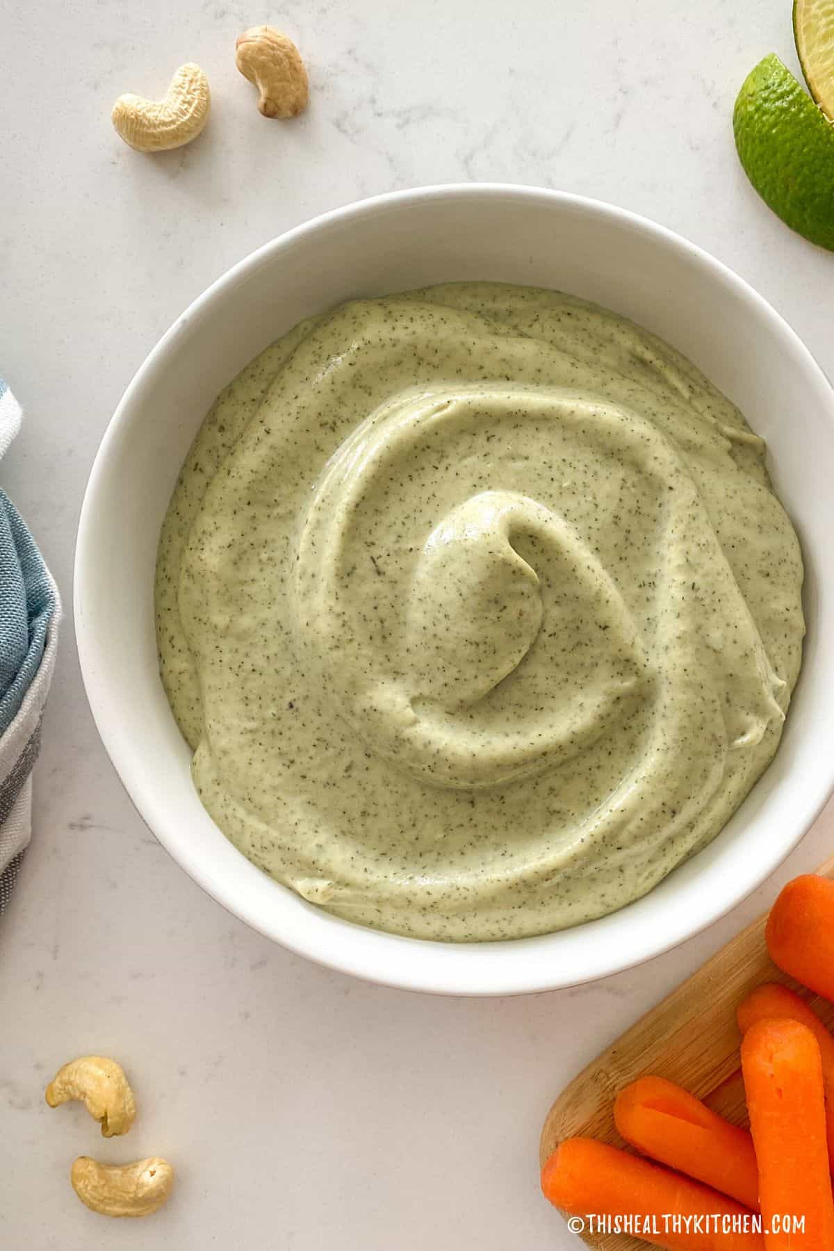 White bowl of cashew ranch dip with carrots and lime wedges on the side.