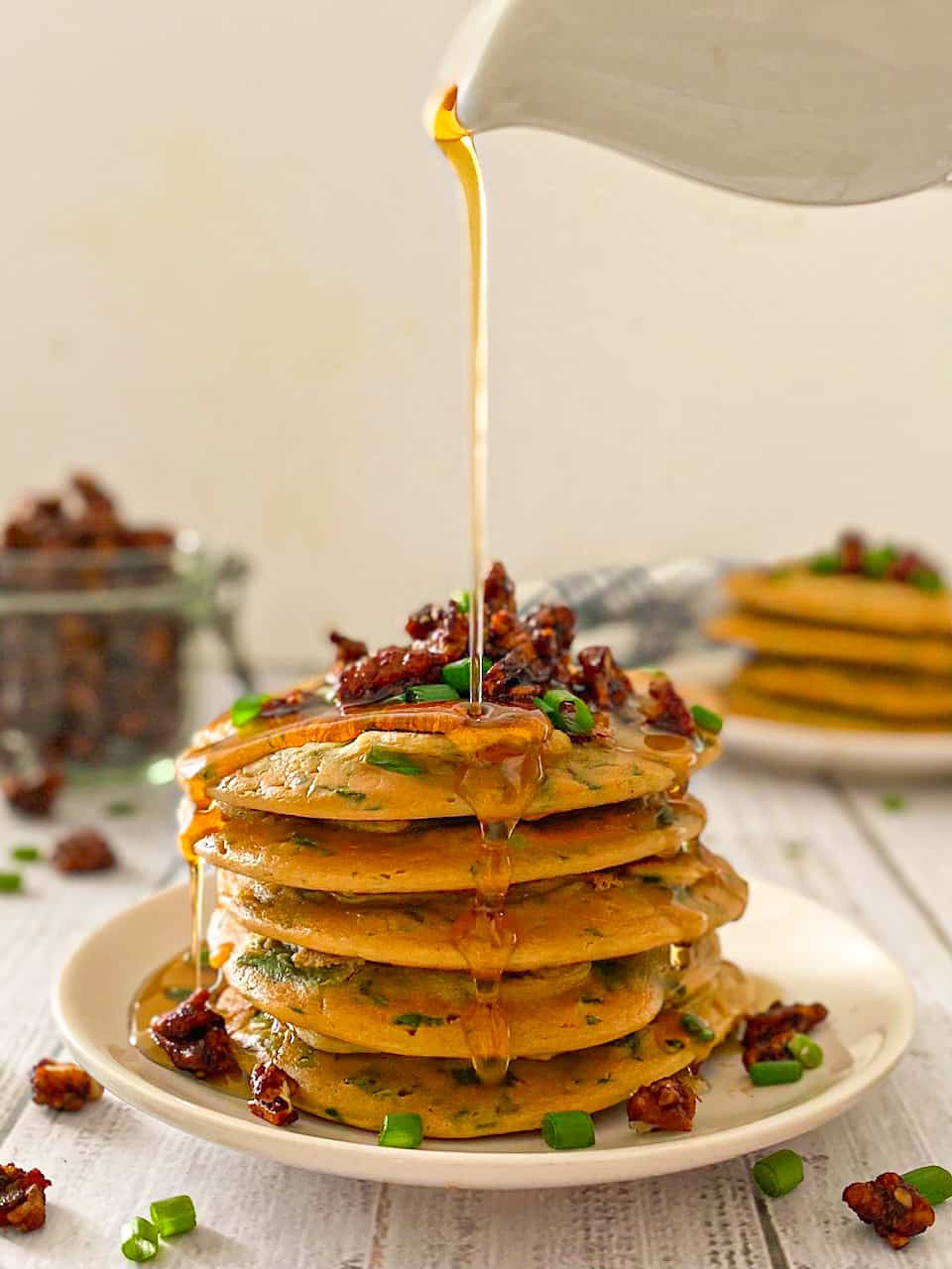 Stack of pancakes with bacon and chives on top and syrup dripping down.