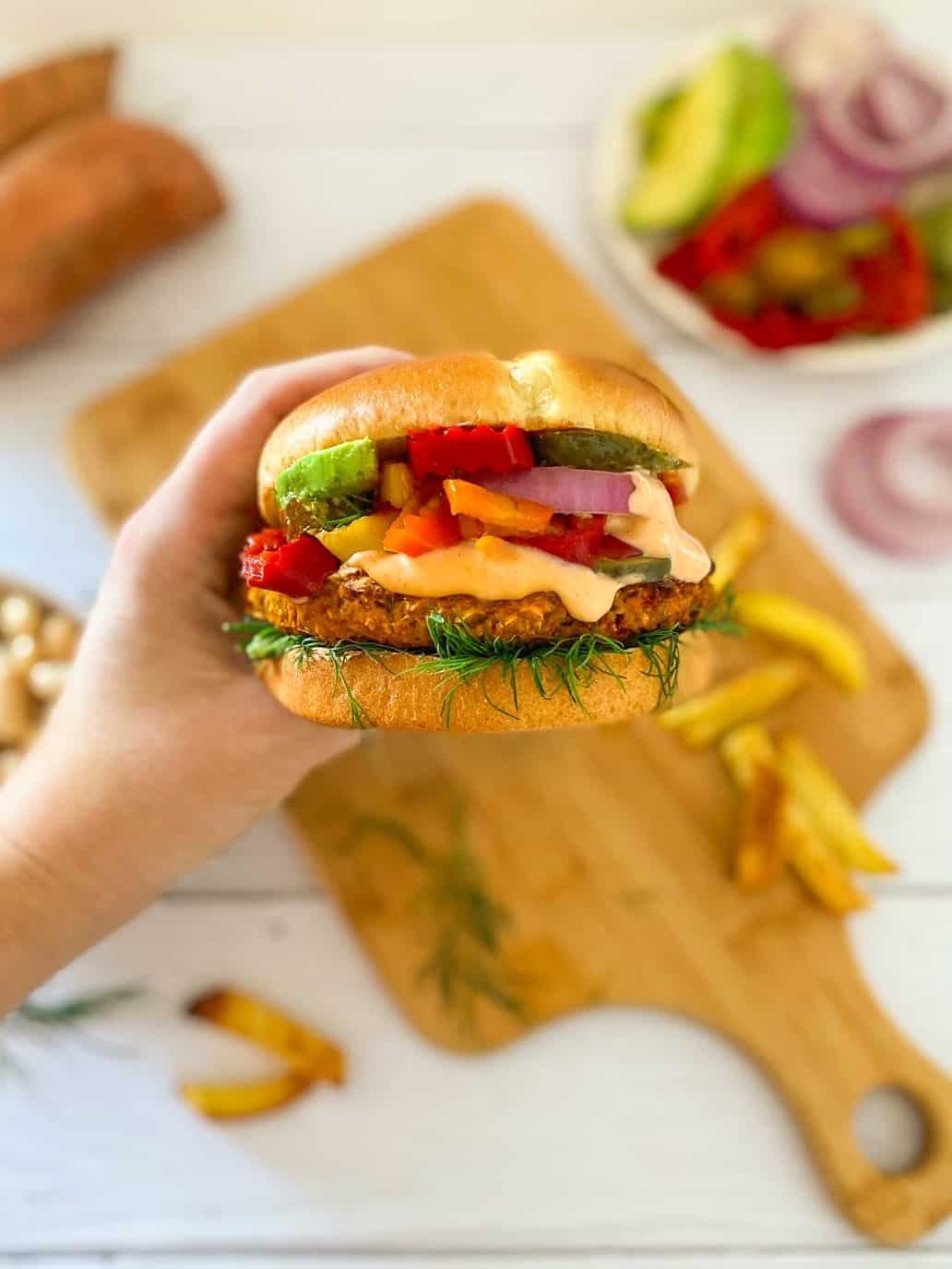 Hand holding vegan sweet potato burger in bun with onion, avocado, peppers and spicy mayo.