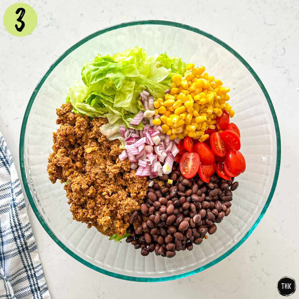 Large glass bowl with vegan taco crumbles, lettuce, corn, red onion, tomato, and black beans inside.