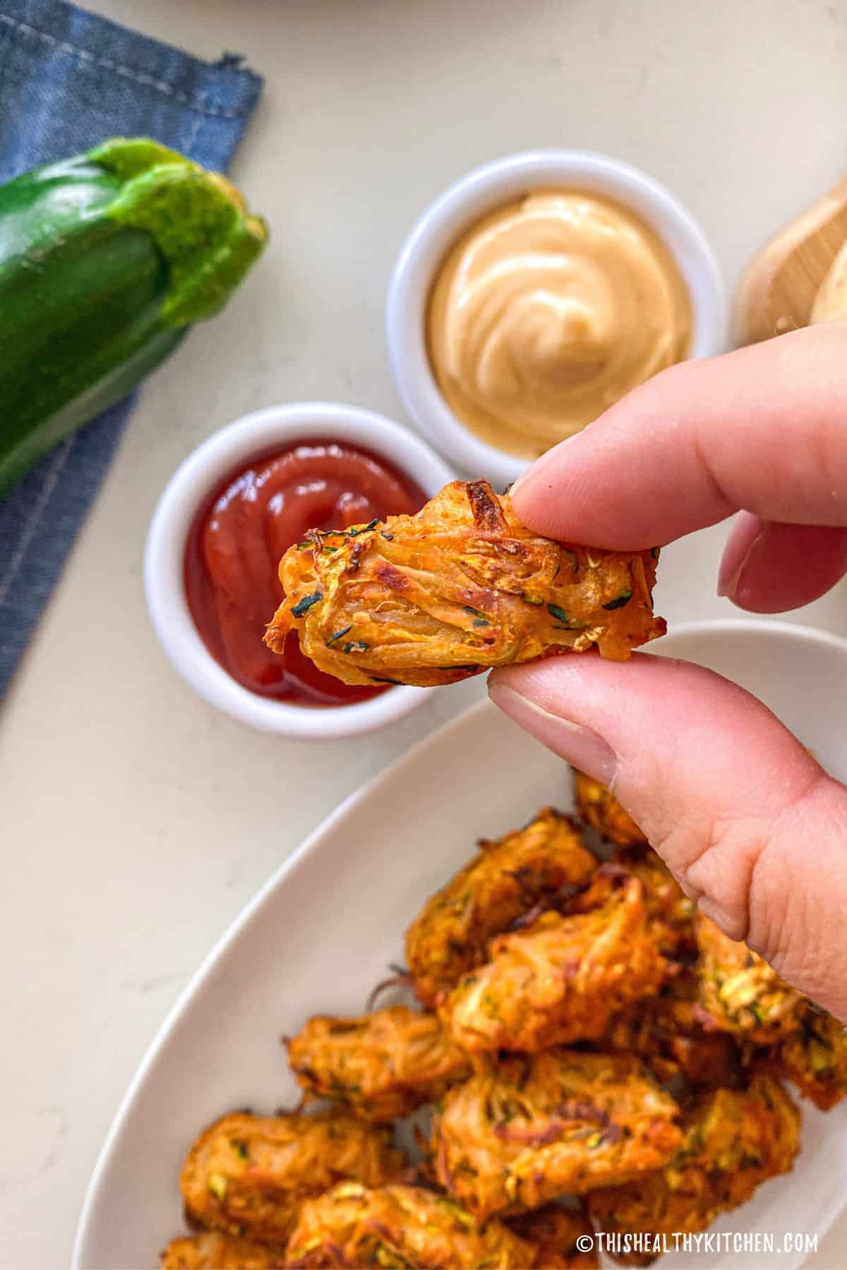 Hand holding up a tater tot, about to dunk into small bowl of ketchup.