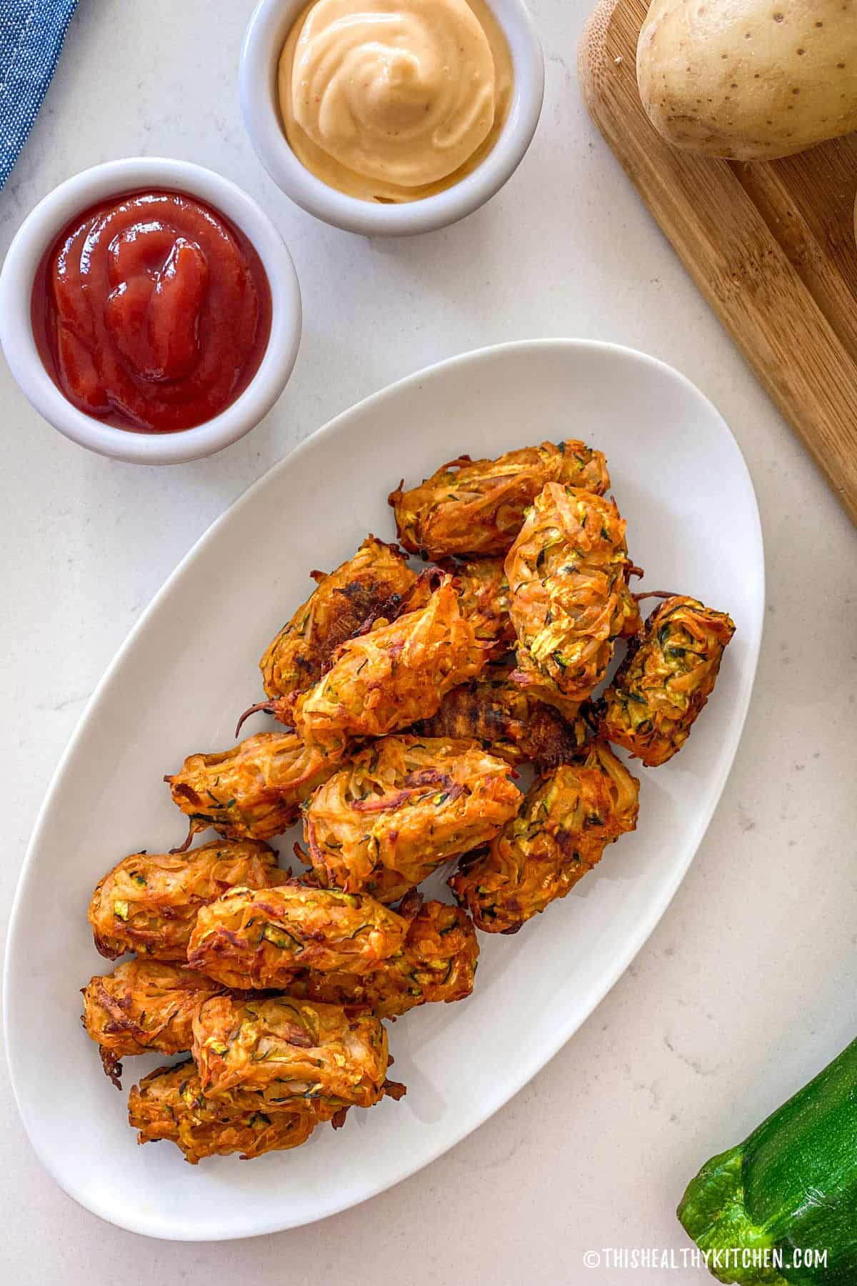 Baked tater tots in white dish with dipping bowls of ketchup and spicy mayo on the side.